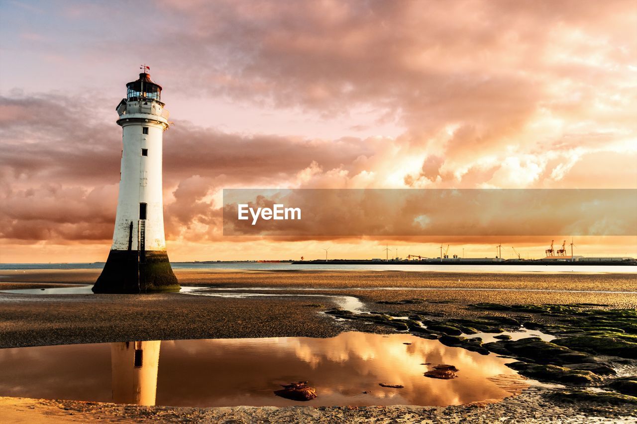 Lighthouse at beach against cloudy sky during sunset