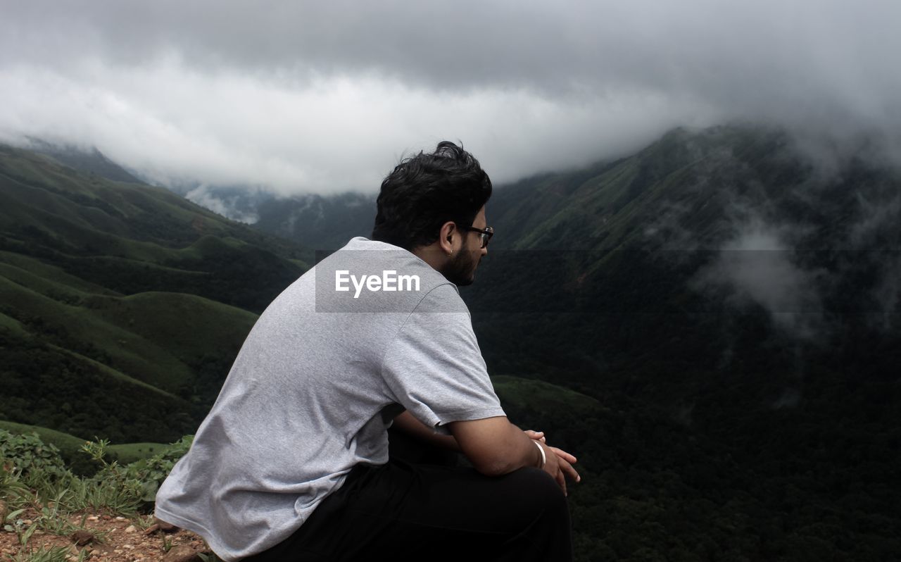 Young man looking at view while sitting on mountain against sky