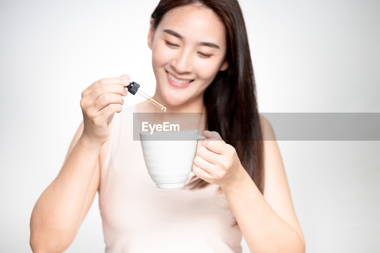 smiling, one person, women, happiness, holding, food and drink, adult, drink, portrait, indoors, studio shot, young adult, refreshment, cheerful, cup, hand, mug, enjoyment, front view, emotion, finger, lifestyles, drinking, white background, smile, teeth, relaxation, wellbeing, food, waist up, hot drink, coffee cup, person, positive emotion, photo shoot, human face, long hair, skin, looking at camera, freshness, coffee, female, casual clothing, hairstyle, tea, brown hair, cut out, copy space, milk, joy, cute, headshot, eating, portrait photography, vitality