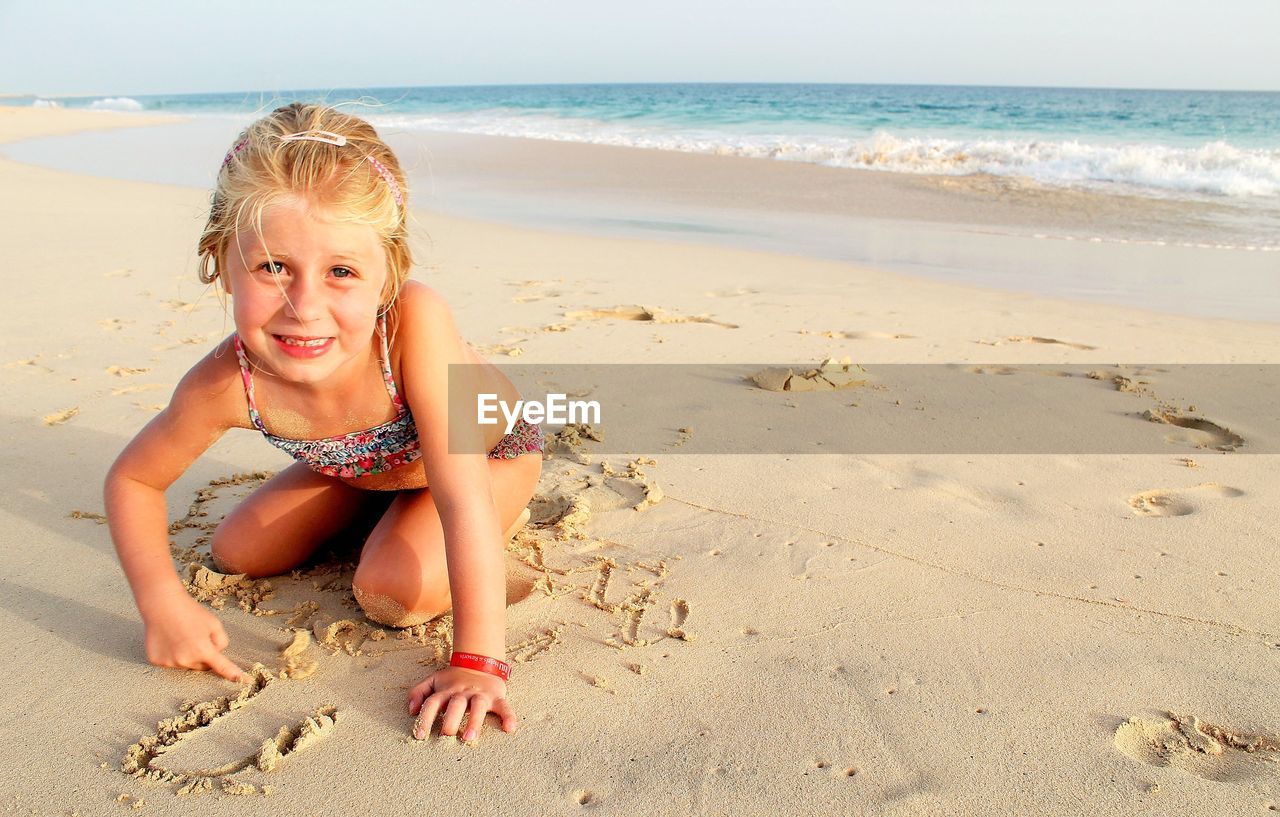 Portrait of smiling girl kneeling at beach against sky
