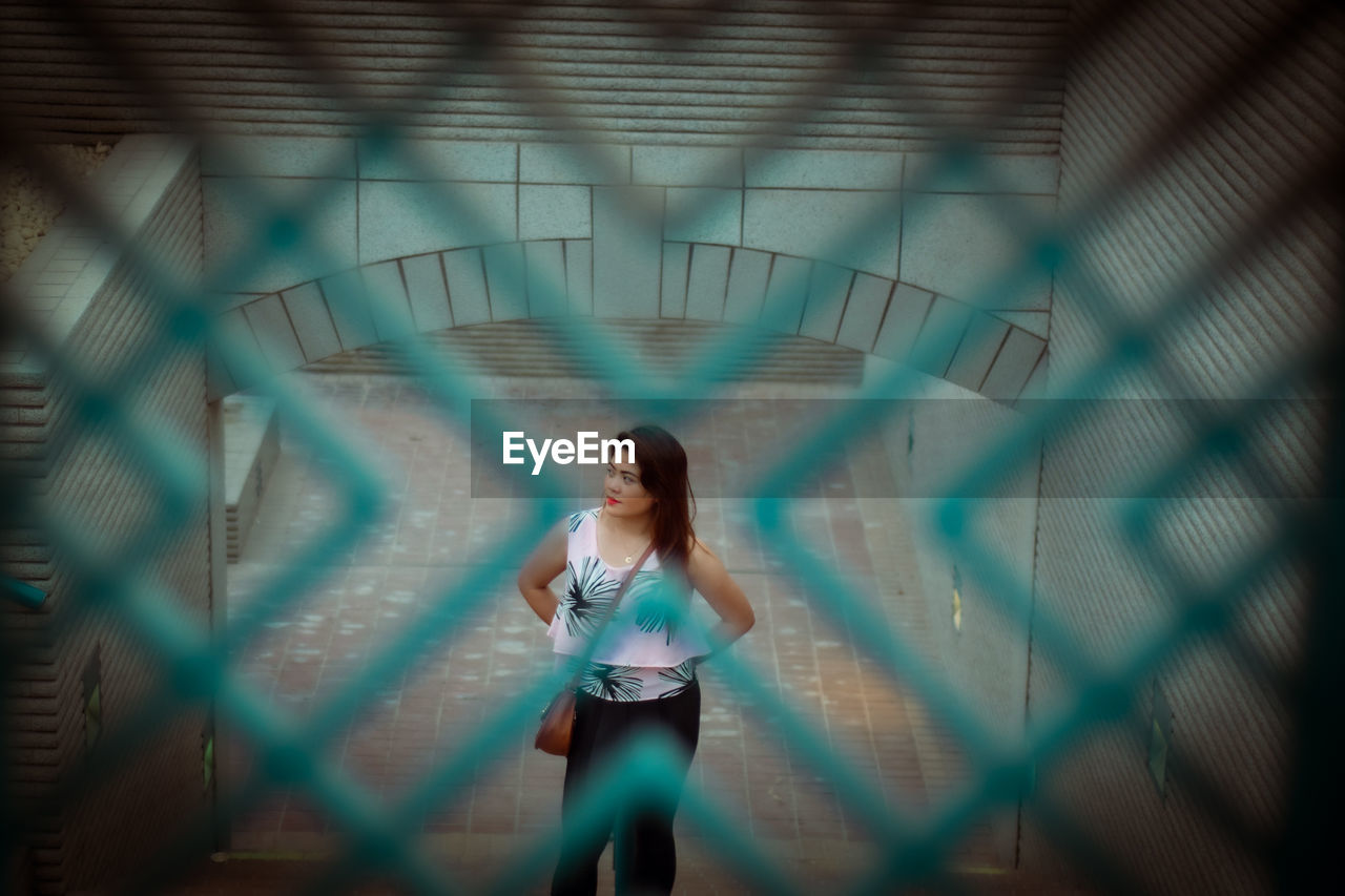 Woman seen through fence looking away while standing on footpath