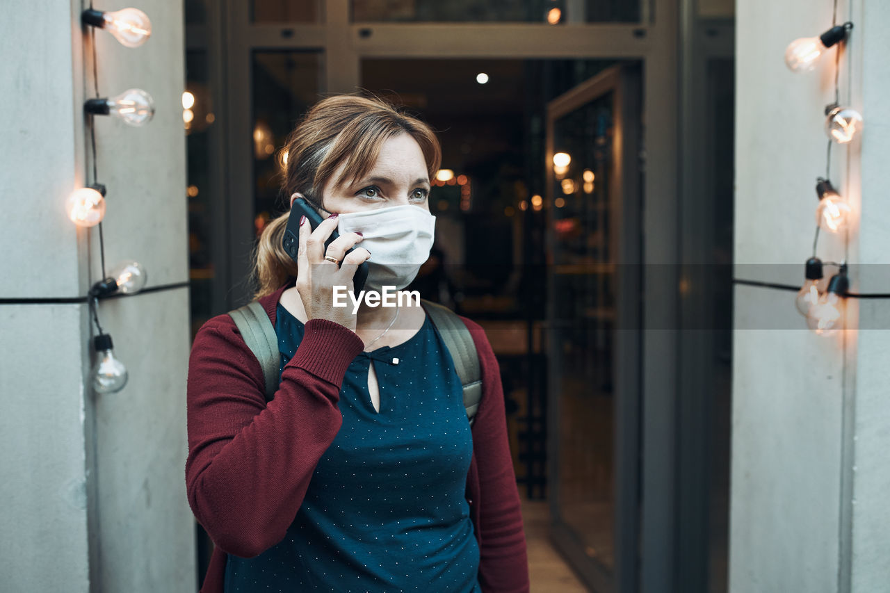 Young woman talking on phone standing in a front of restaurant downtown wearing the face mask