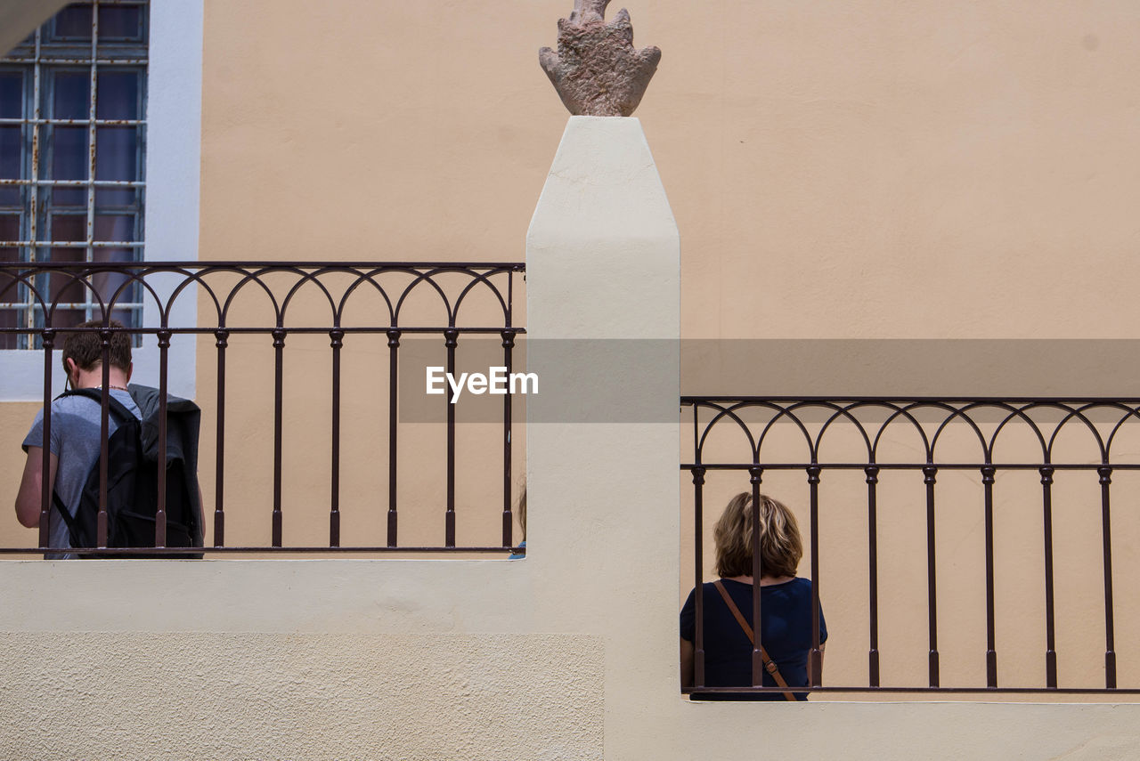 REAR VIEW OF PEOPLE SITTING ON RAILING AGAINST BUILDING