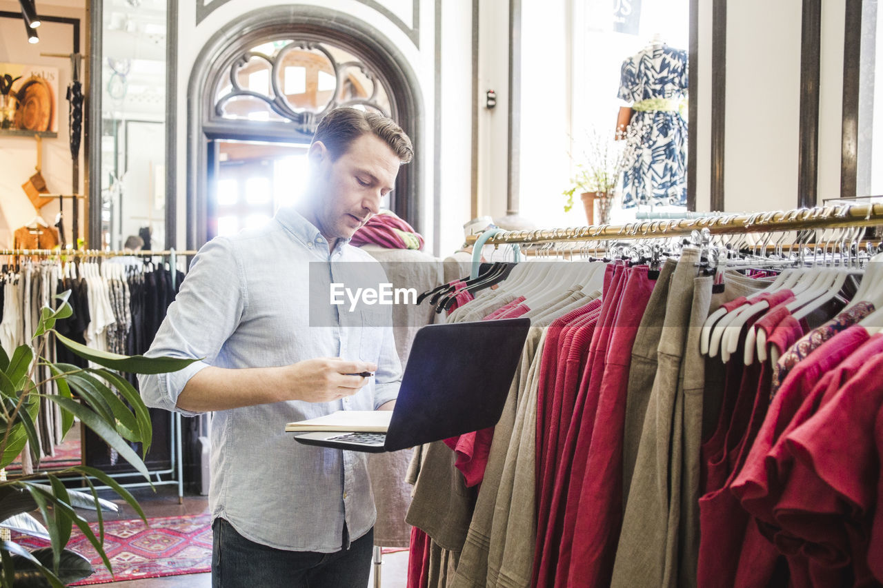 Confident male entrepreneur using laptop while standing by clothes rack in boutique