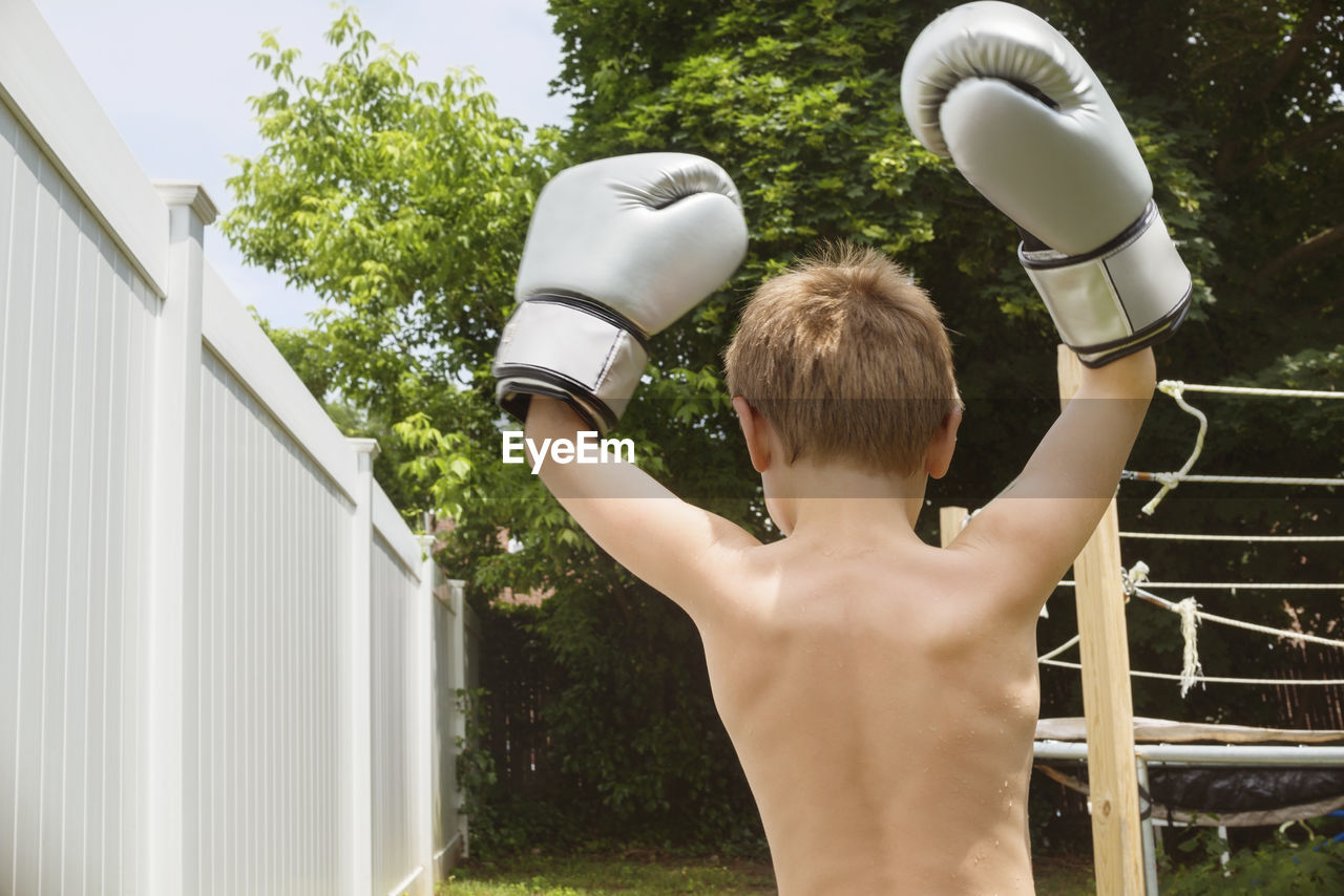 Rear view of boy raising arms while wearing boxing gloves at backyard