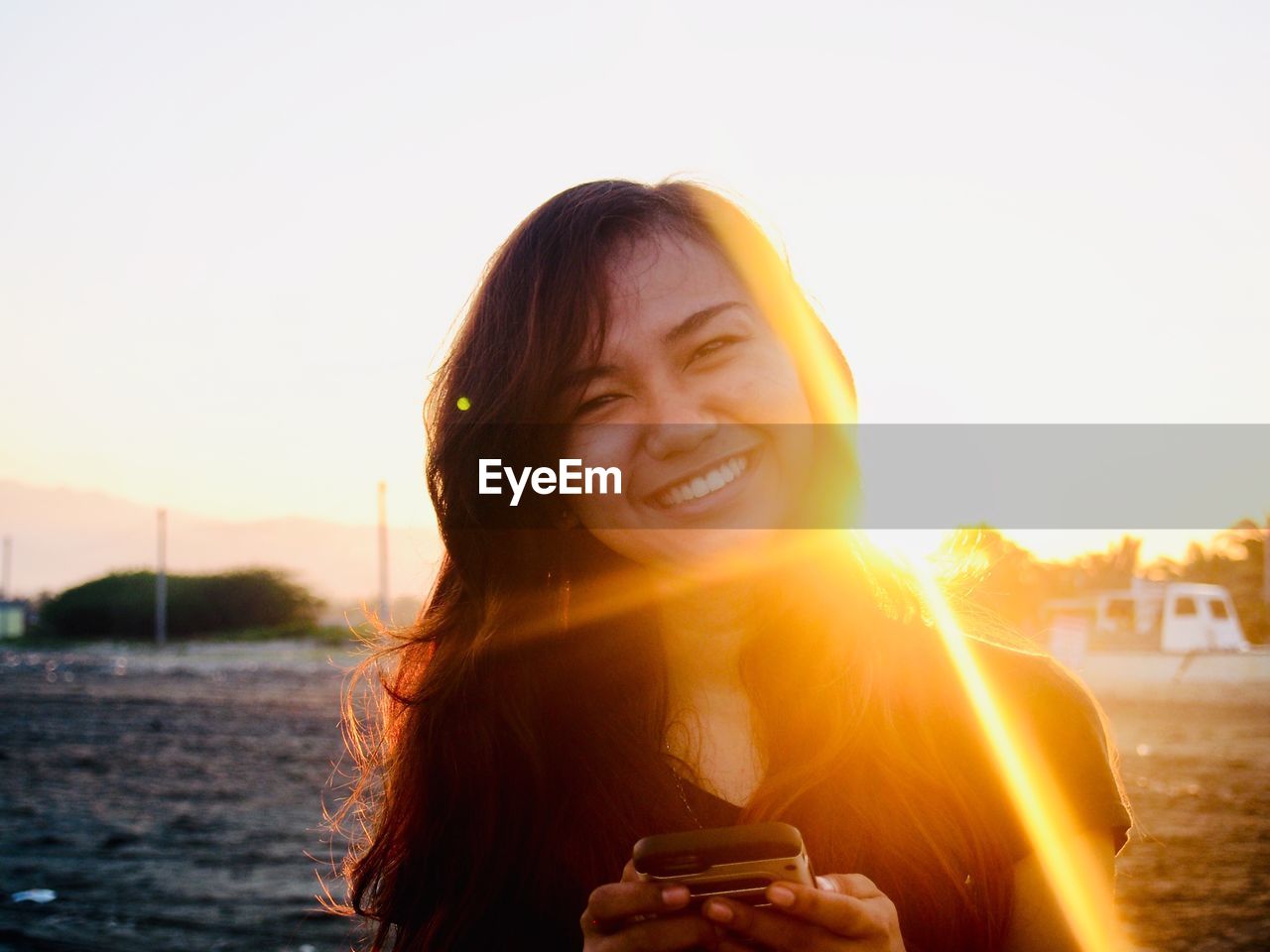 Portrait of happy young woman using smart phone against clear sky during sunset