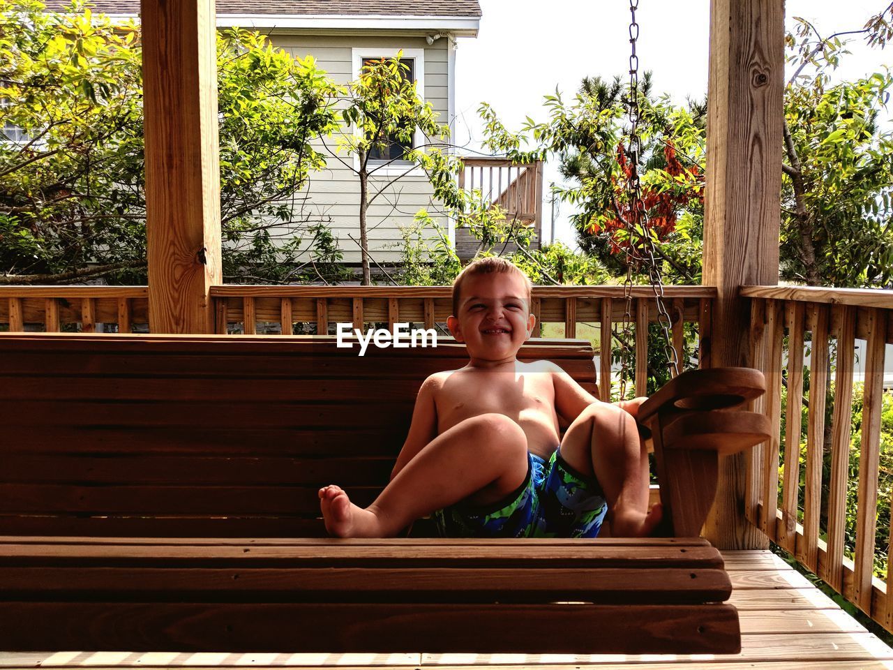 Portrait of shirtless boy smiling while sitting on swing at porch