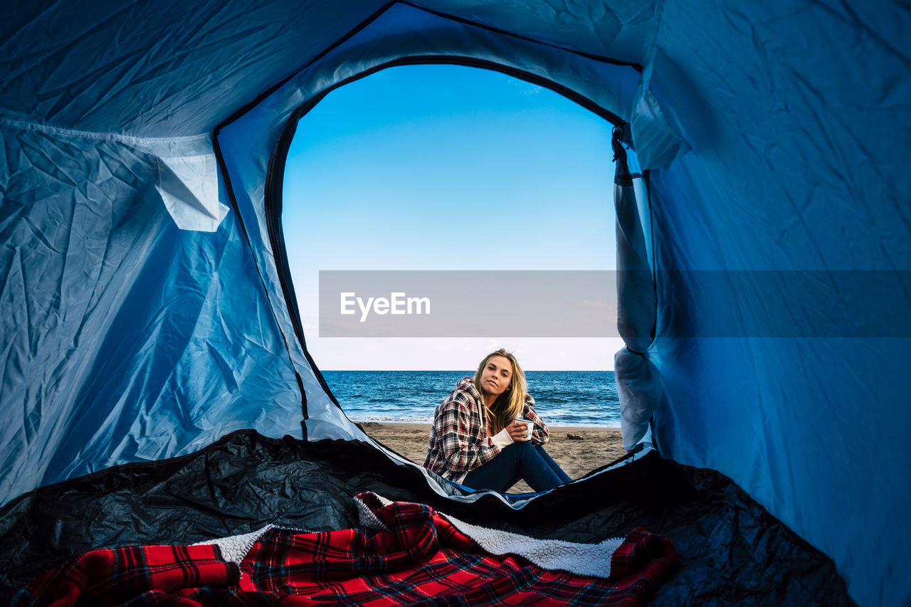 Portrait of woman sitting by tent at beach against sky