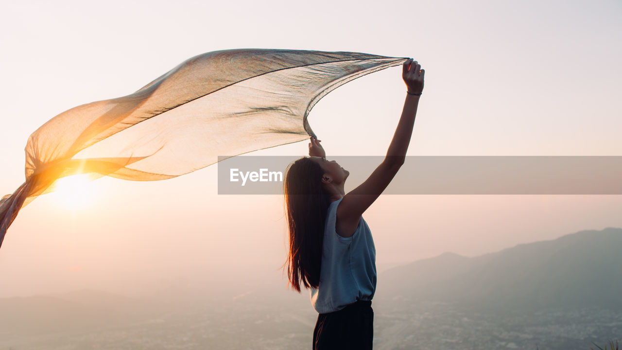 Side view of woman holding sarong against sky during sunset