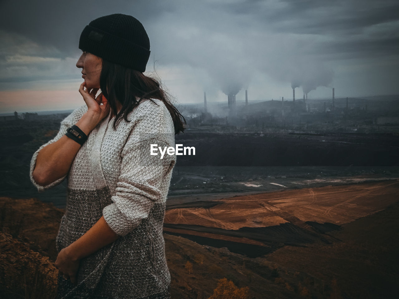 Thoughtful woman looking away while standing against factory emitting smoke