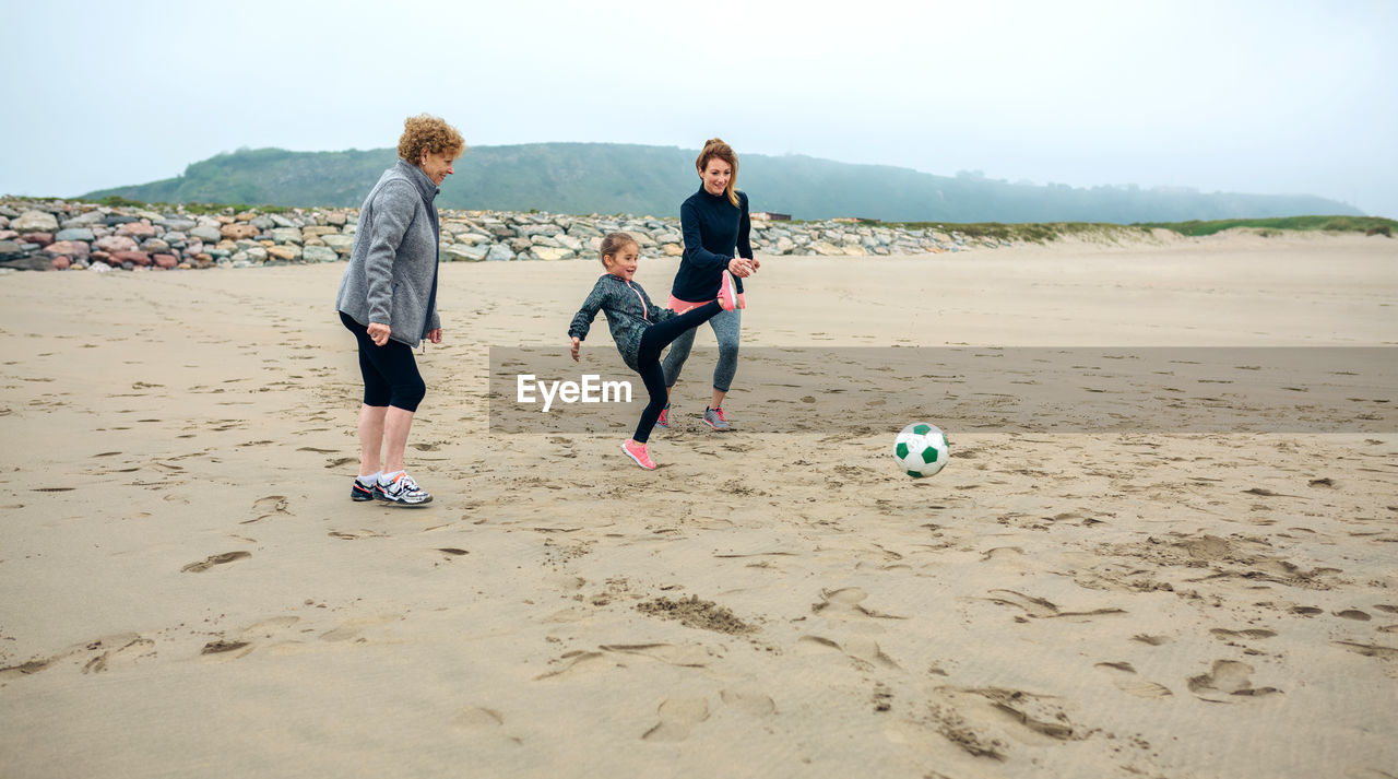 Woman with mother and daughter playing ball at beach against sky