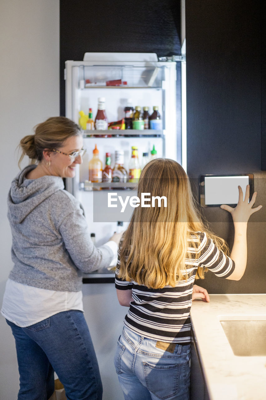 Daughter using digital tablet while mother looking standing by refrigerator at smart home