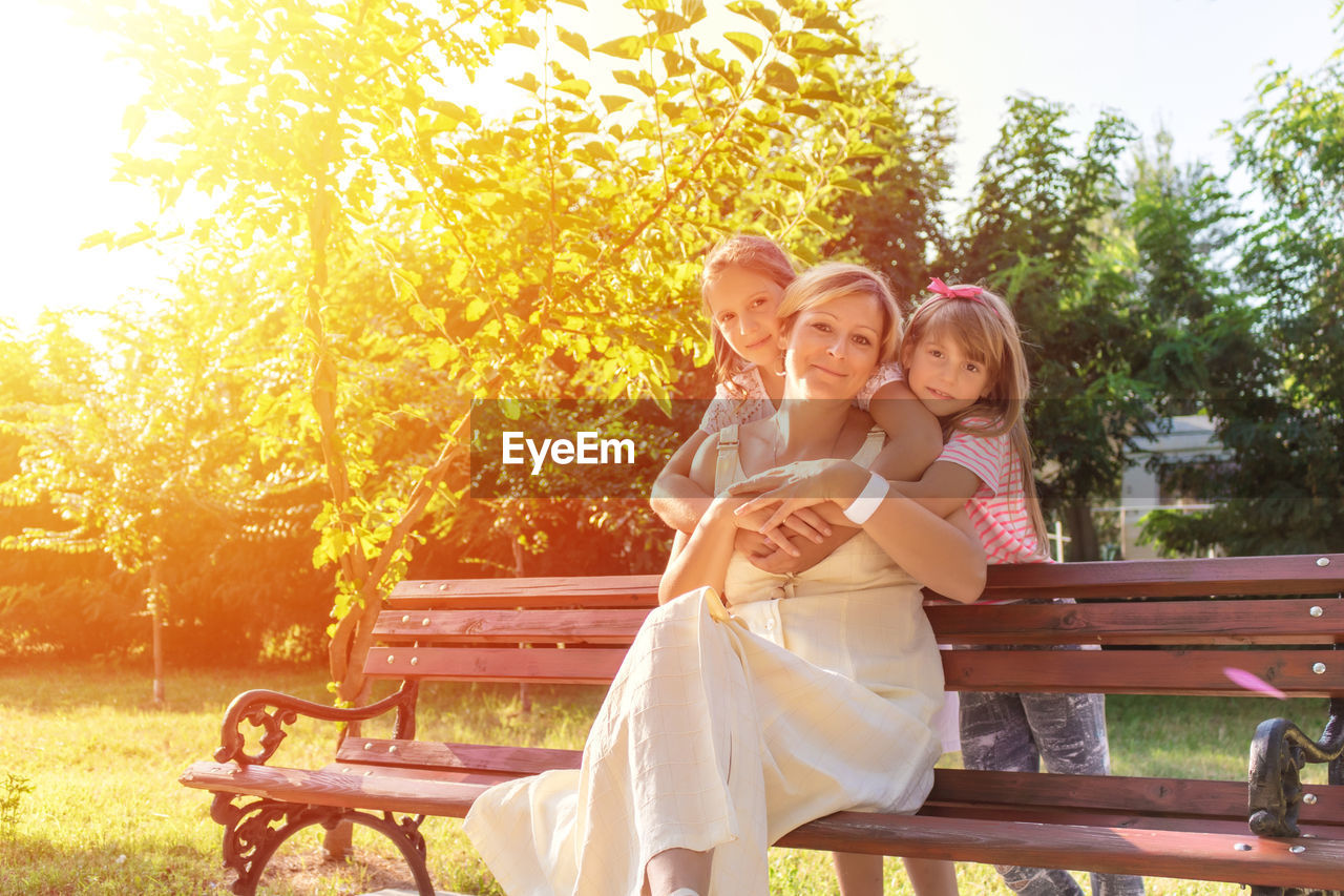 Portrait of mother sitting on bench with daughters trees in park