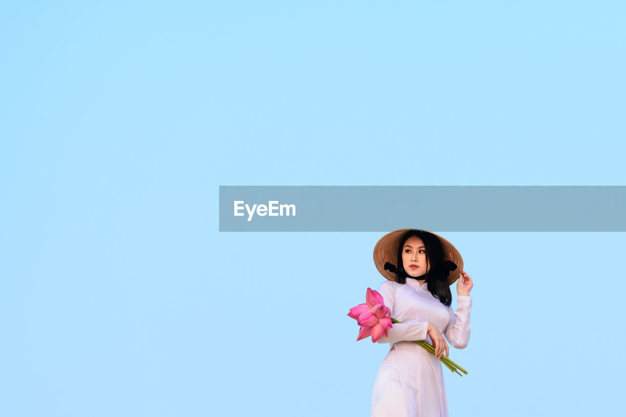 Beautiful young woman holding lotus water lilies while standing against clear blue sky