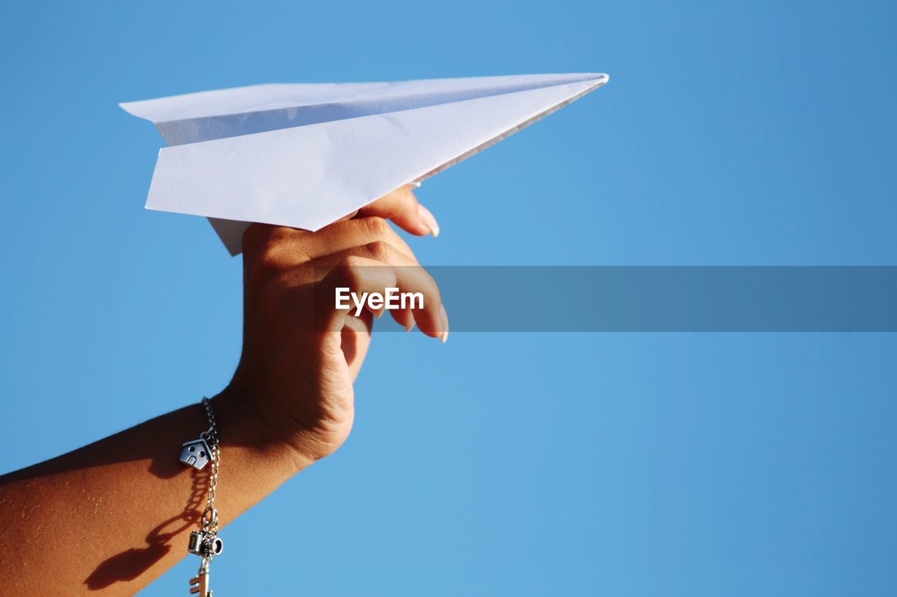 Cropped hand of woman holding paper airplane against clear blue sky