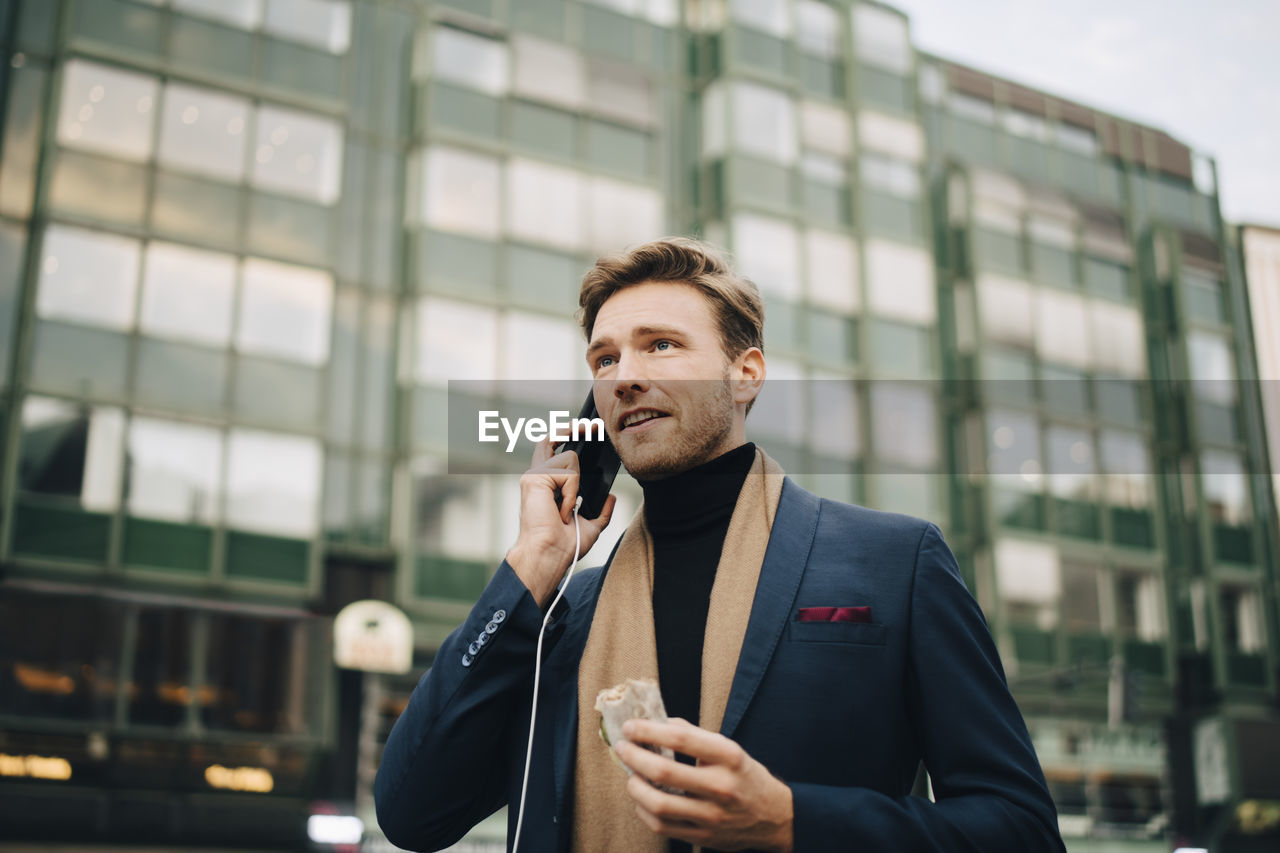 Confident businessman with wrap sandwich talking through smart phone while standing in city