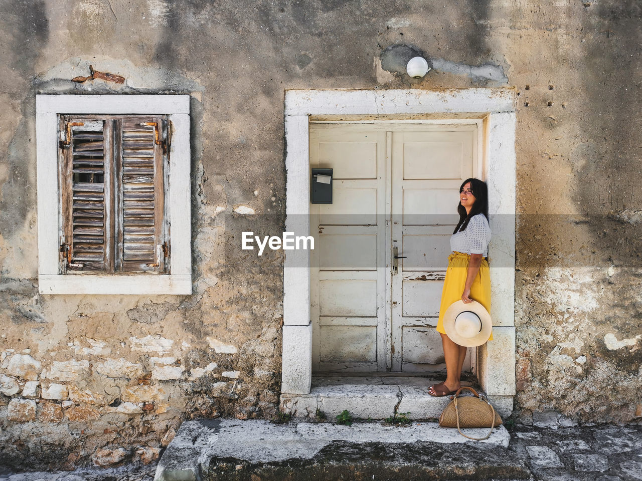 Young woman in summer outfit standing in front of old white wooden door of an old house.