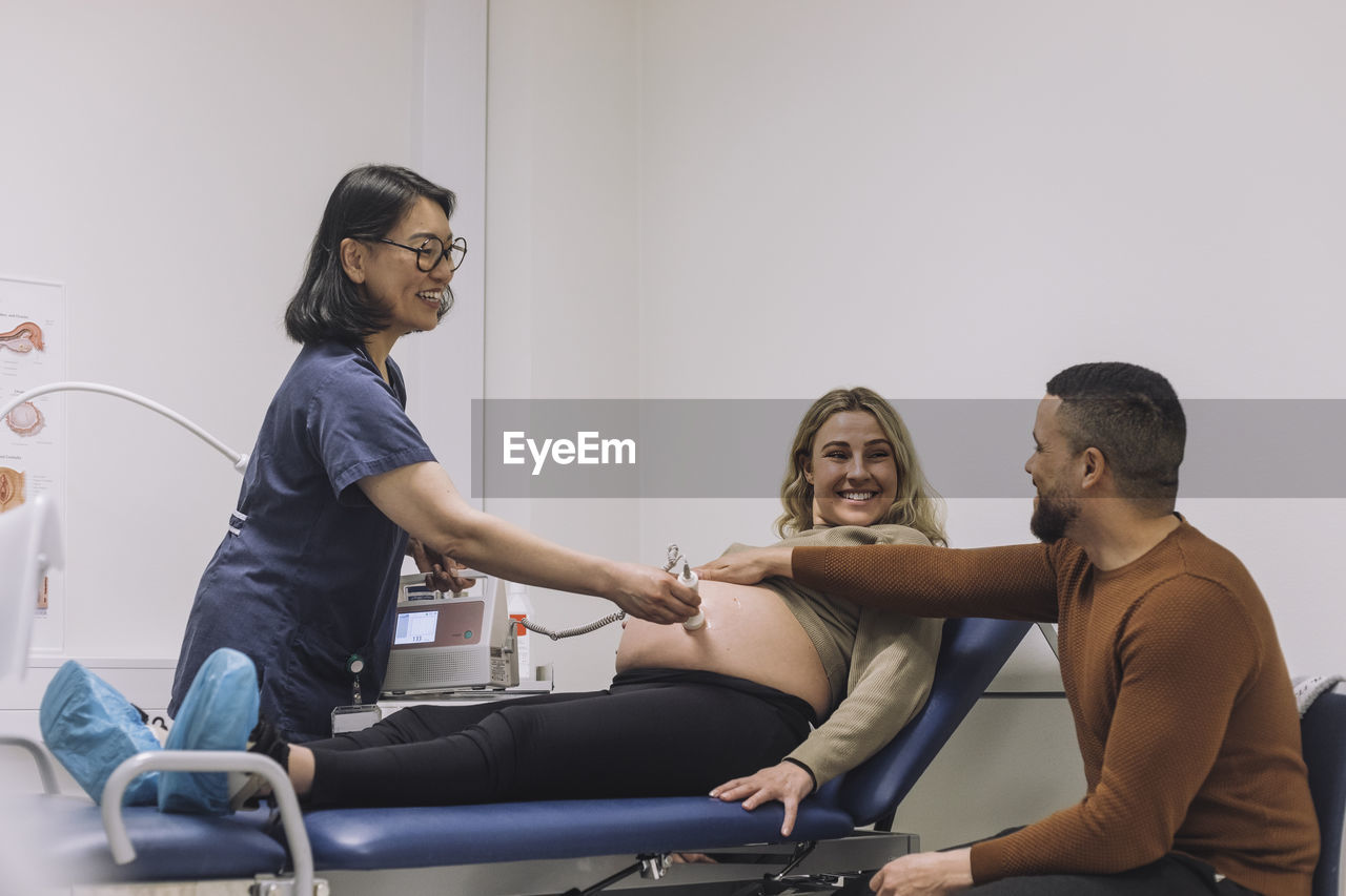 Happy gynecologist doing ultrasound looking at man touching stomach of pregnant woman in hospital