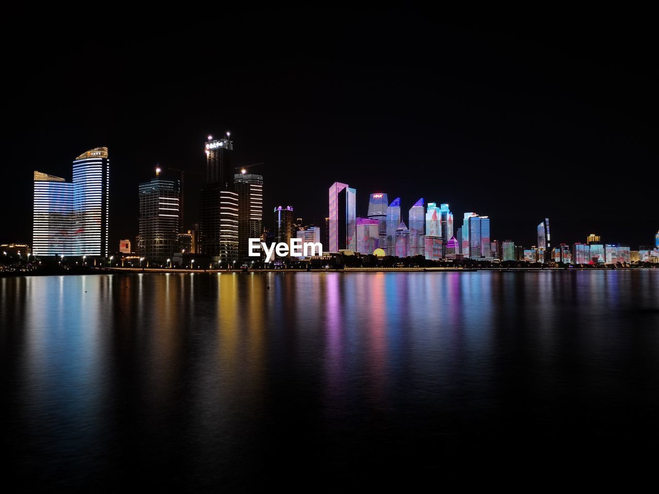 Illuminated modern buildings by river against sky at night