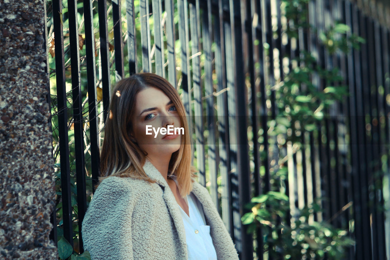 Portrait of young woman standing by fence at park