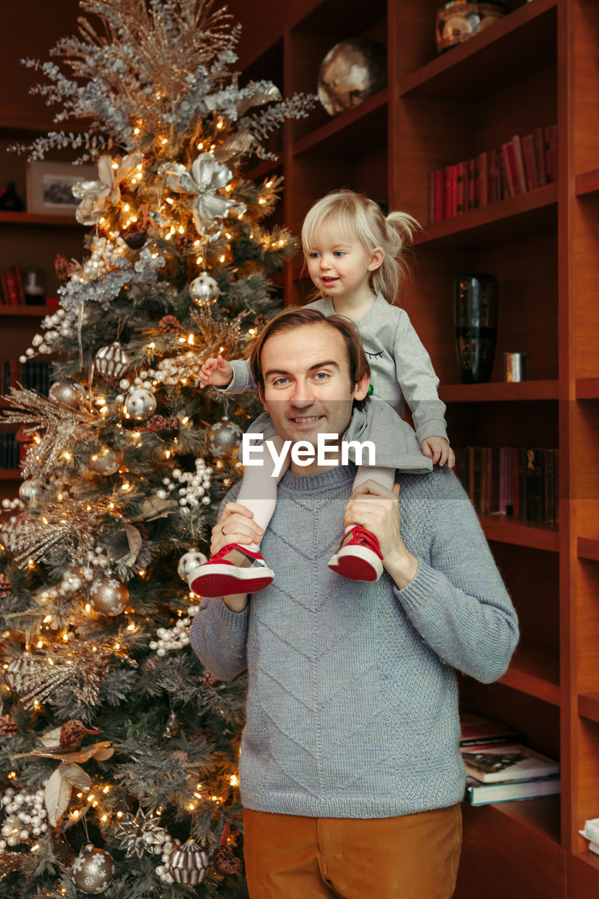 Smiling caucasian father holding baby girl on shoulders by decorated christmas tree. happy family 