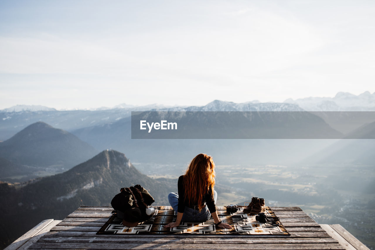 Back view of unrecognizable female tourist sitting at viewpoint in mountains and observing amazing scenery in morning