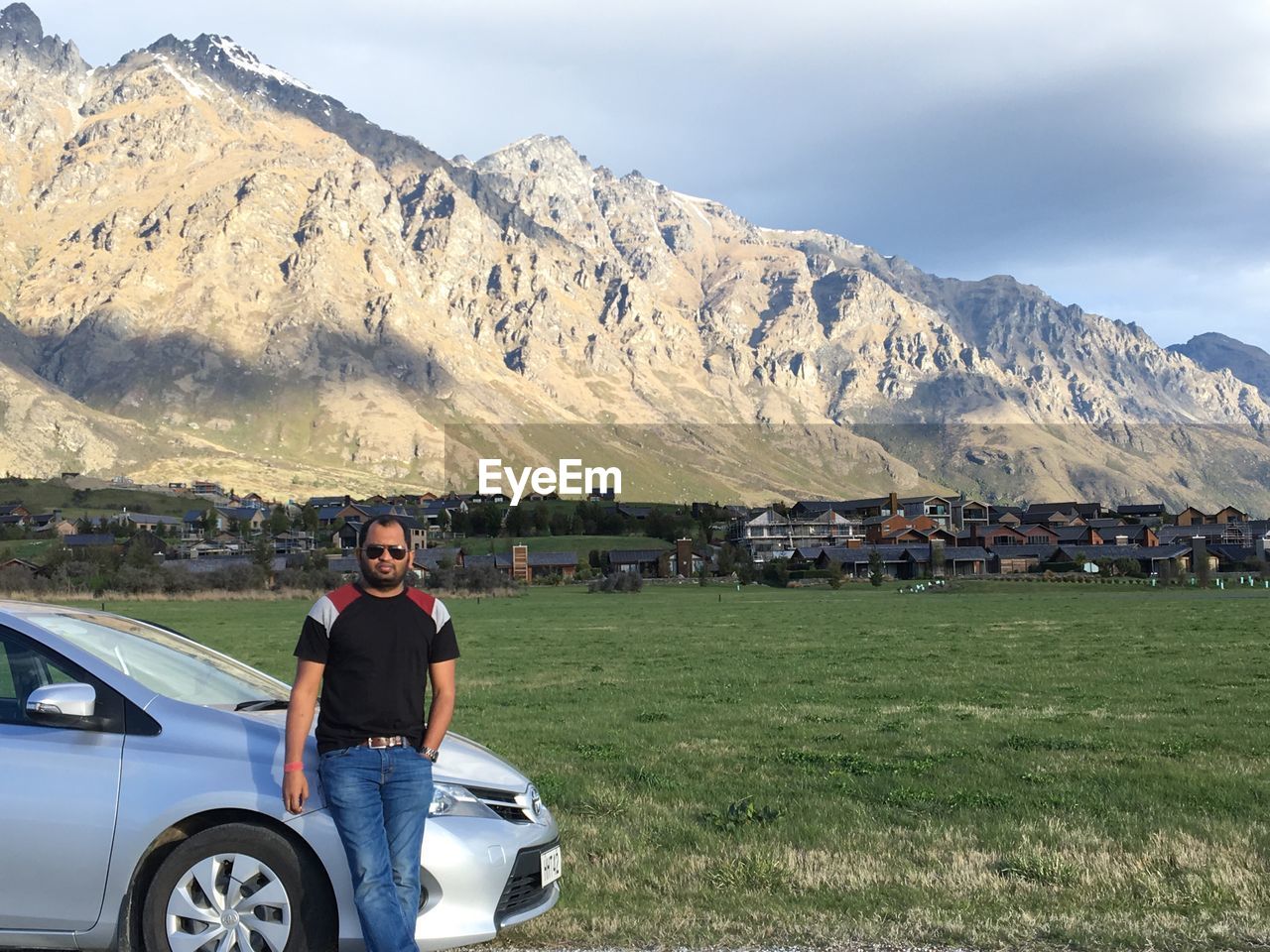 PORTRAIT OF MAN STANDING BY CAR ON FIELD AGAINST MOUNTAIN