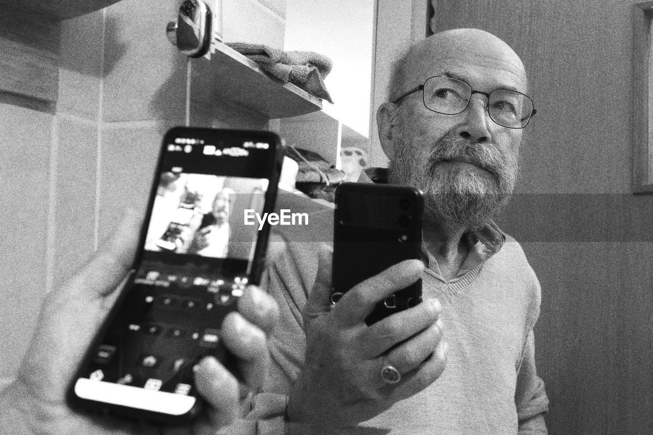 adult, senior adult, technology, black and white, men, person, one person, holding, facial hair, seniors, indoors, eyeglasses, glasses, wireless technology, monochrome photography, portrait, human face, lifestyles, beard, communication, monochrome, portable information device, smartphone, activity, mobile phone, black, waist up, selfie