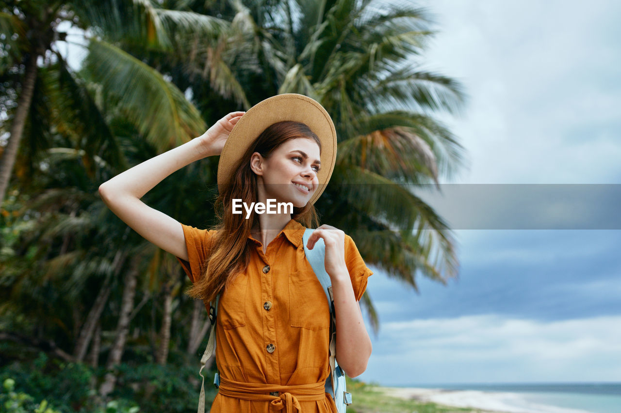 YOUNG WOMAN LOOKING AWAY WHILE STANDING BY TREE