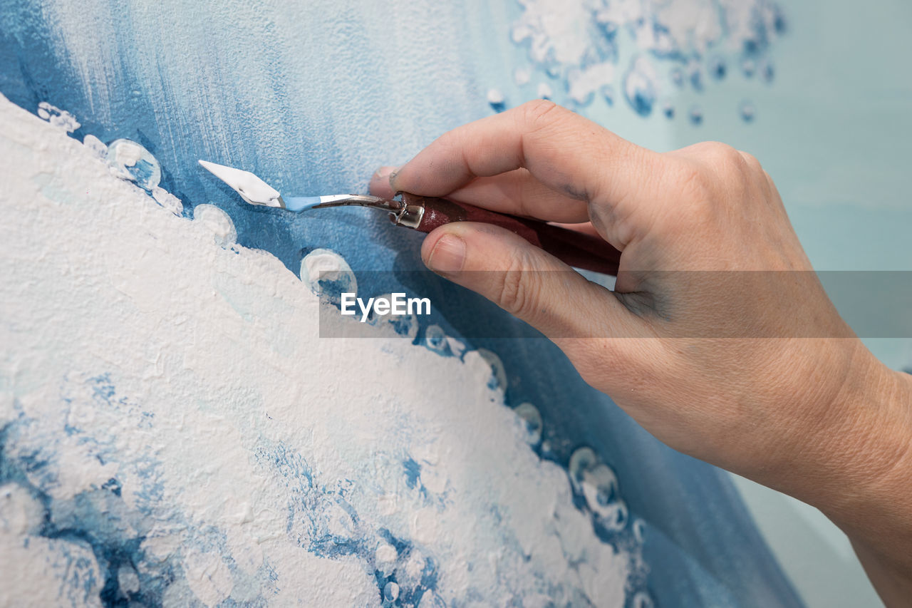 The hand of a woman artist draws an image of a sea wave using a palette knife, close uo