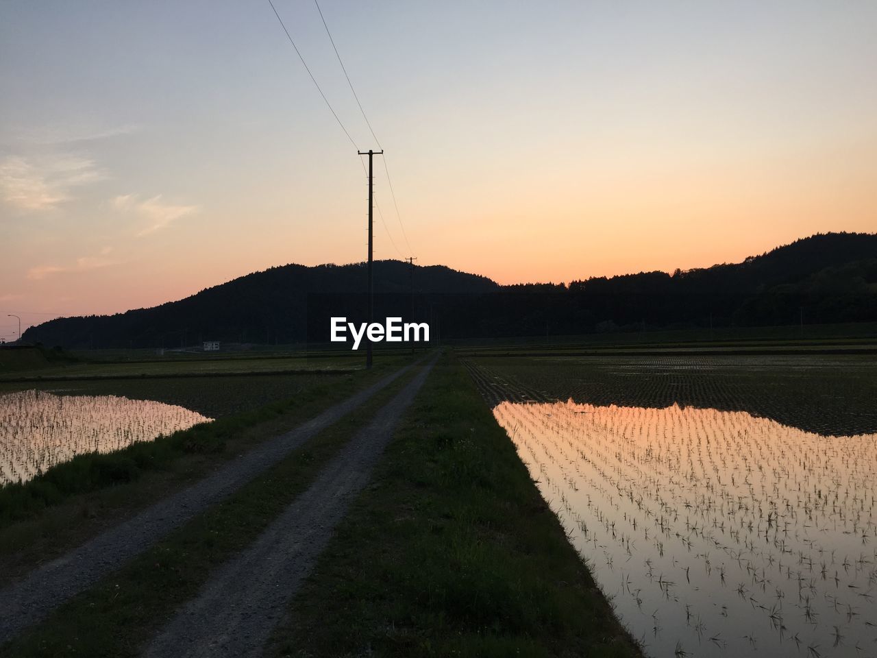 Dirt road amidst field in front of mountains against sky during sunset