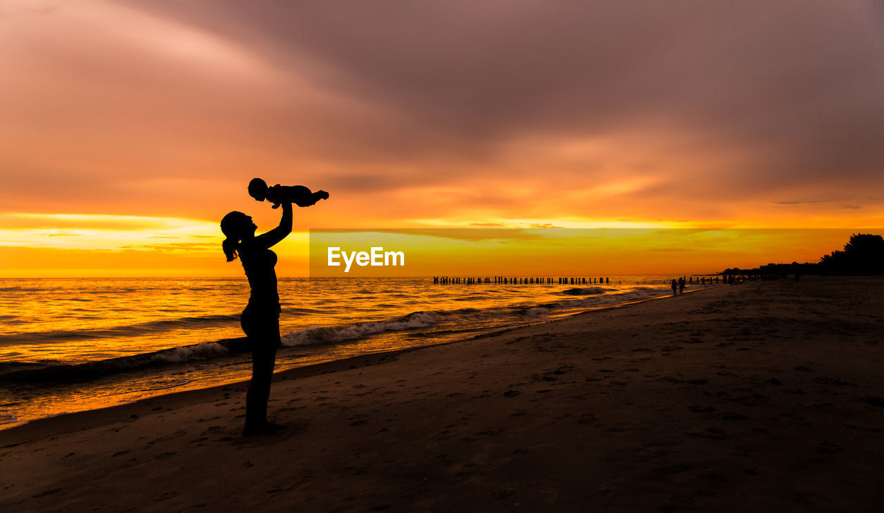 Young adult woman mother holding baby in air on beach with sunset background silhouette
