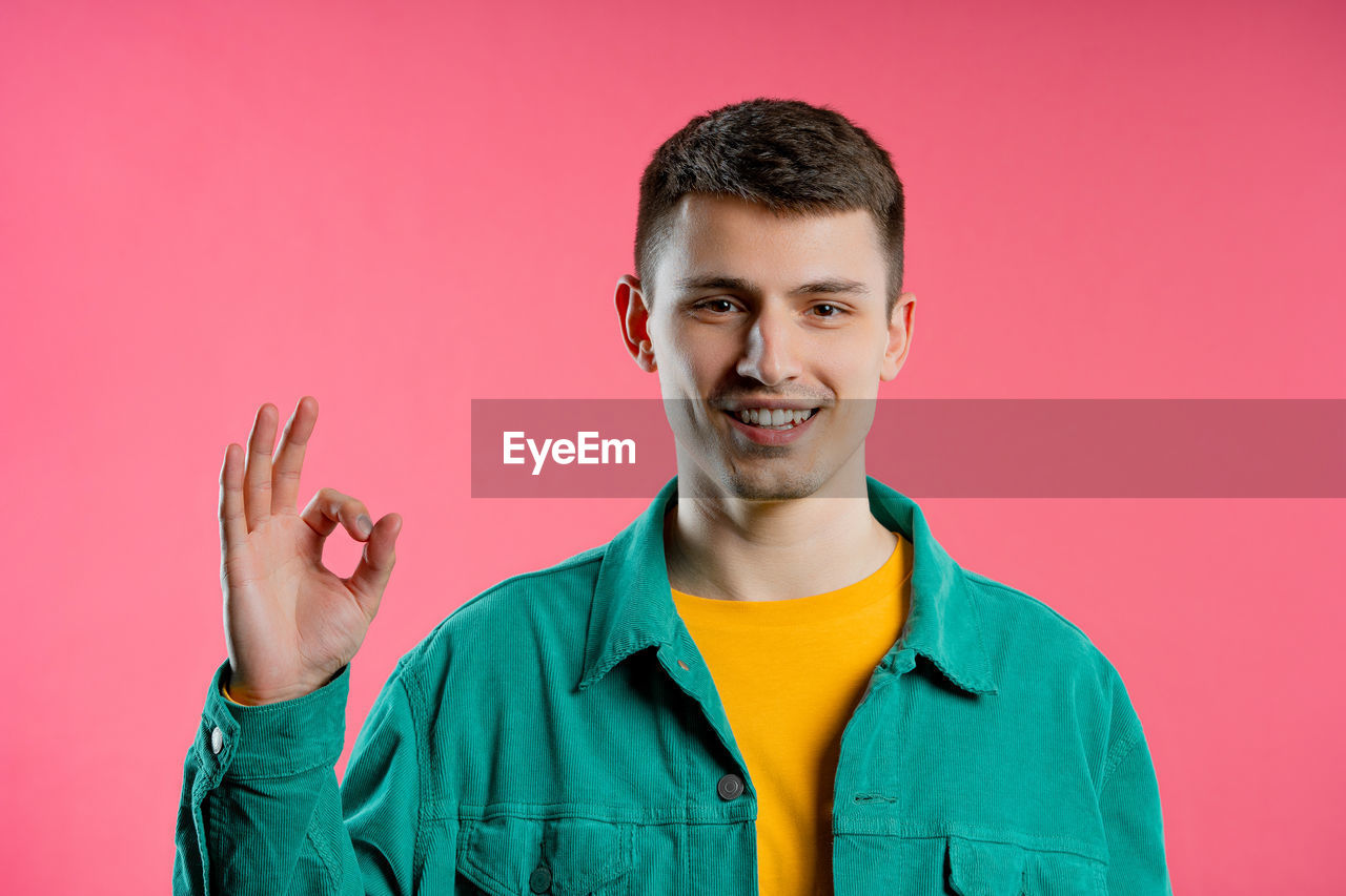 portrait, smiling, colored background, studio shot, one person, happiness, young adult, looking at camera, emotion, cheerful, indoors, men, adult, front view, person, positive emotion, teeth, waist up, pink background, headshot, smile, casual clothing, pink, sign language, clothing, singing, teenager, standing, copy space, fun, gesturing