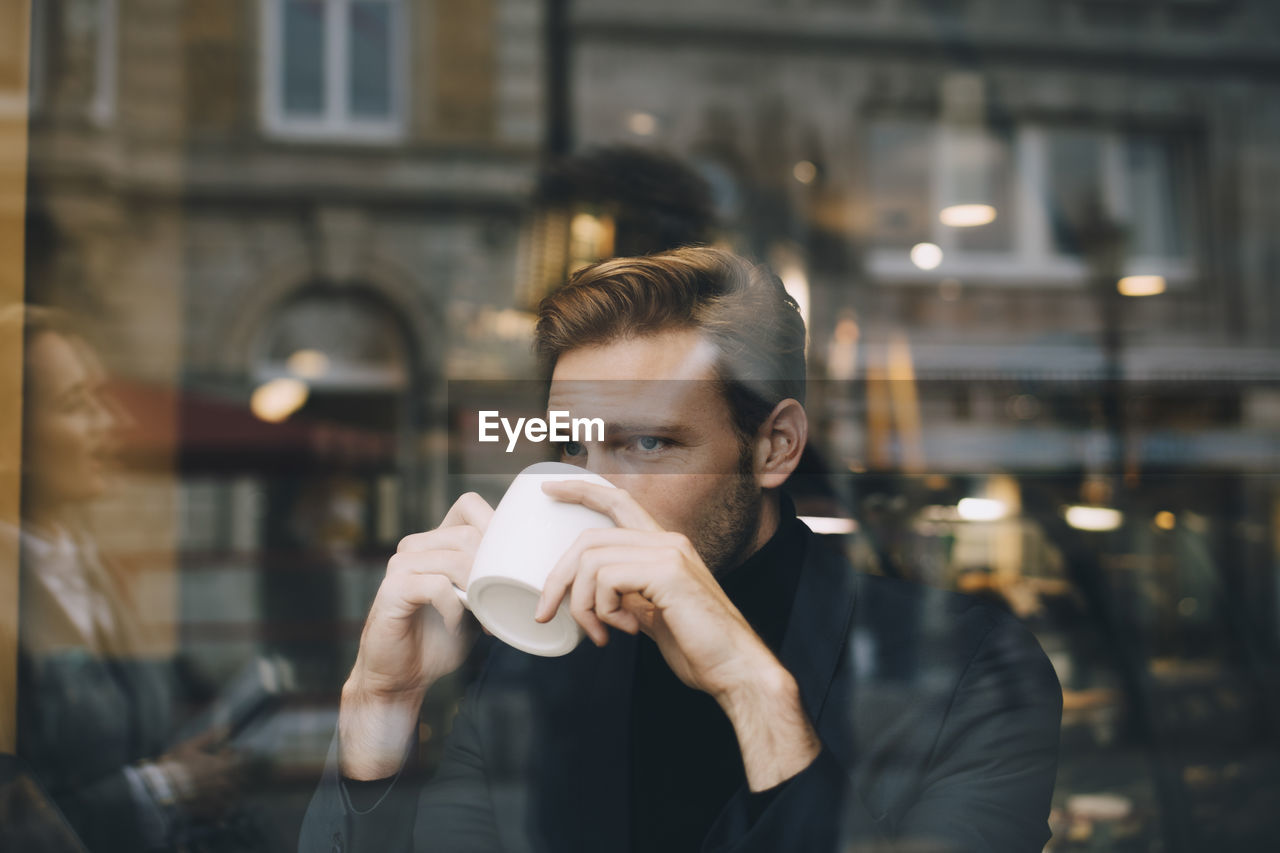 Businessman drinking coffee while looking away in cafe seen through glass window