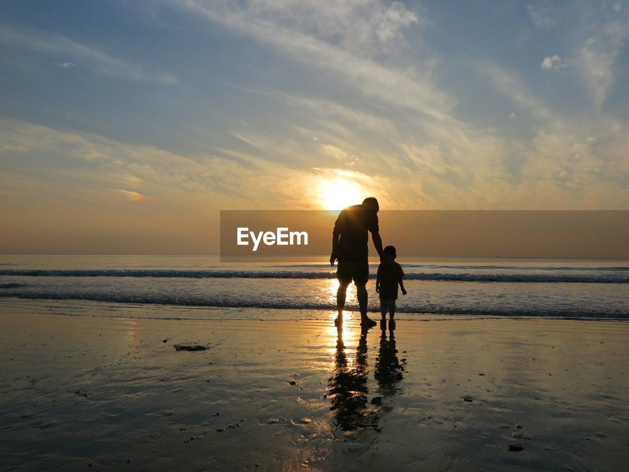 Silhouette rear view of man and son on wet shore at beach against sky during sunset