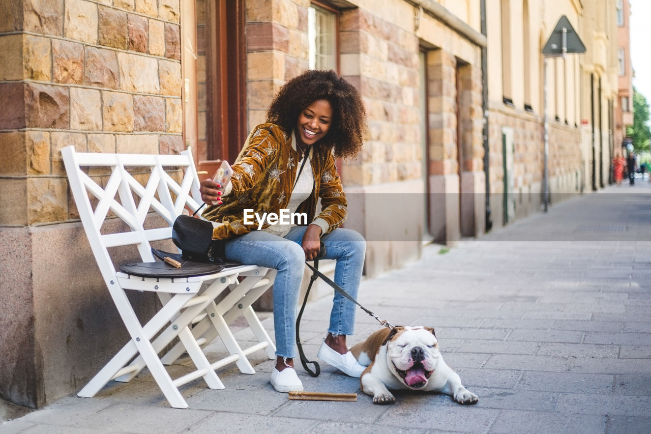 Happy woman sitting with dog taking selfie on mobile phone at sidewalk in city