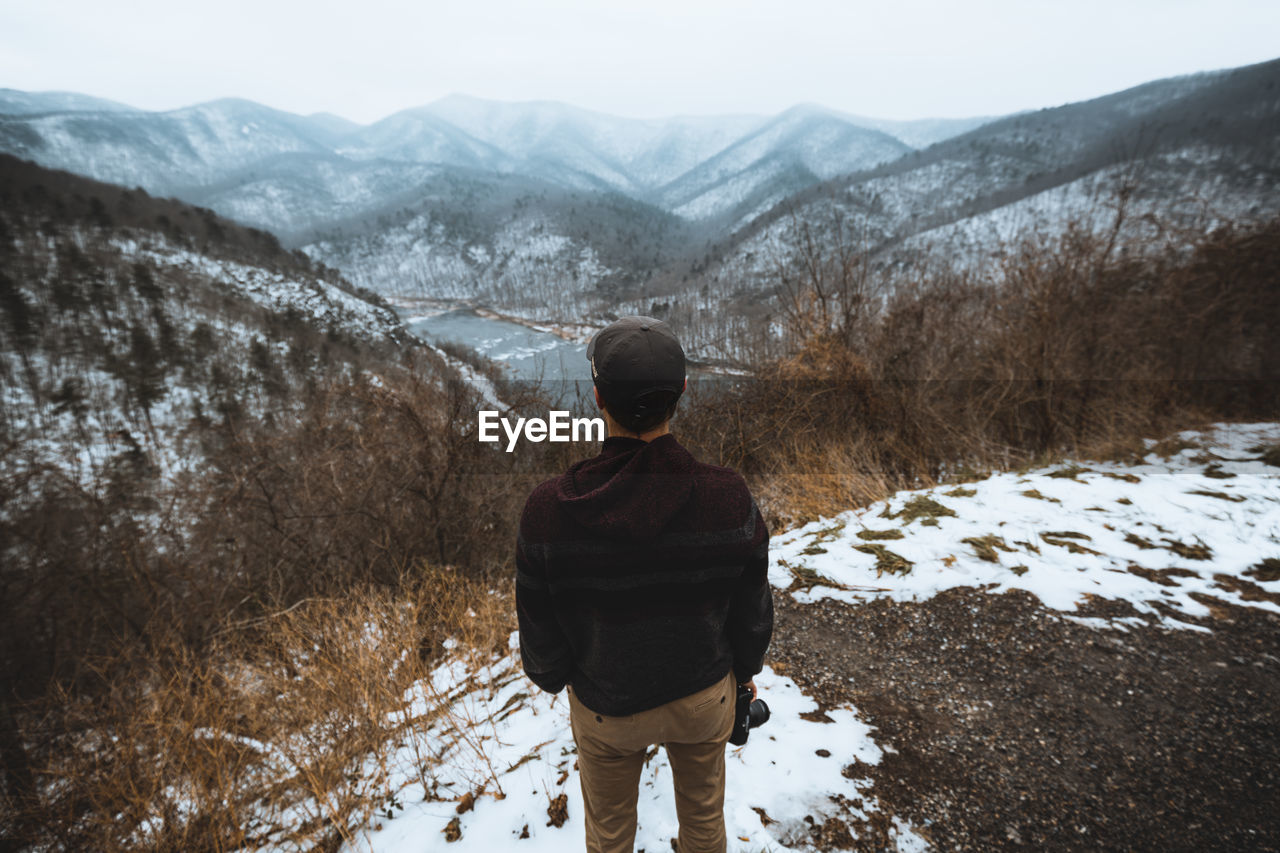 REAR VIEW OF MAN STANDING ON SNOWCAPPED MOUNTAINS