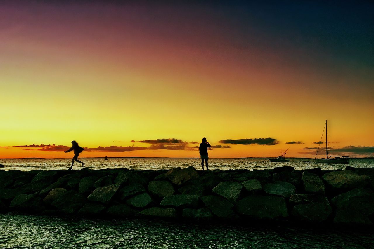 Silhouette people on groyne in sea against orange sunset sky