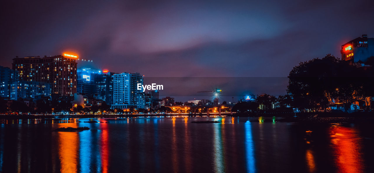 Illuminated buildings by river against sky at night