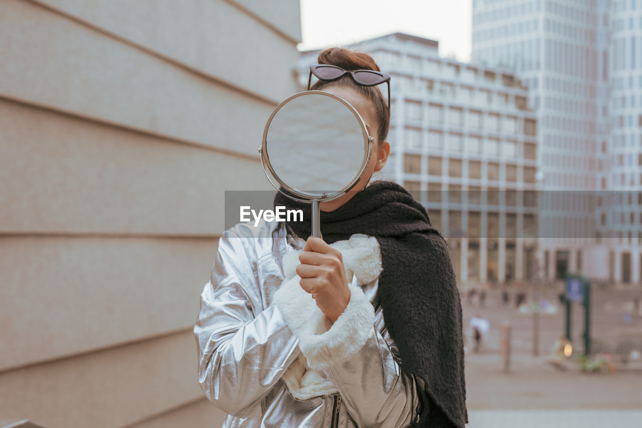 Young woman holding mirror while standing against buildings in city