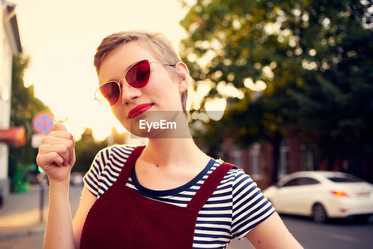 PORTRAIT OF YOUNG WOMAN WEARING SUNGLASSES STANDING AGAINST CITY IN BACKGROUND