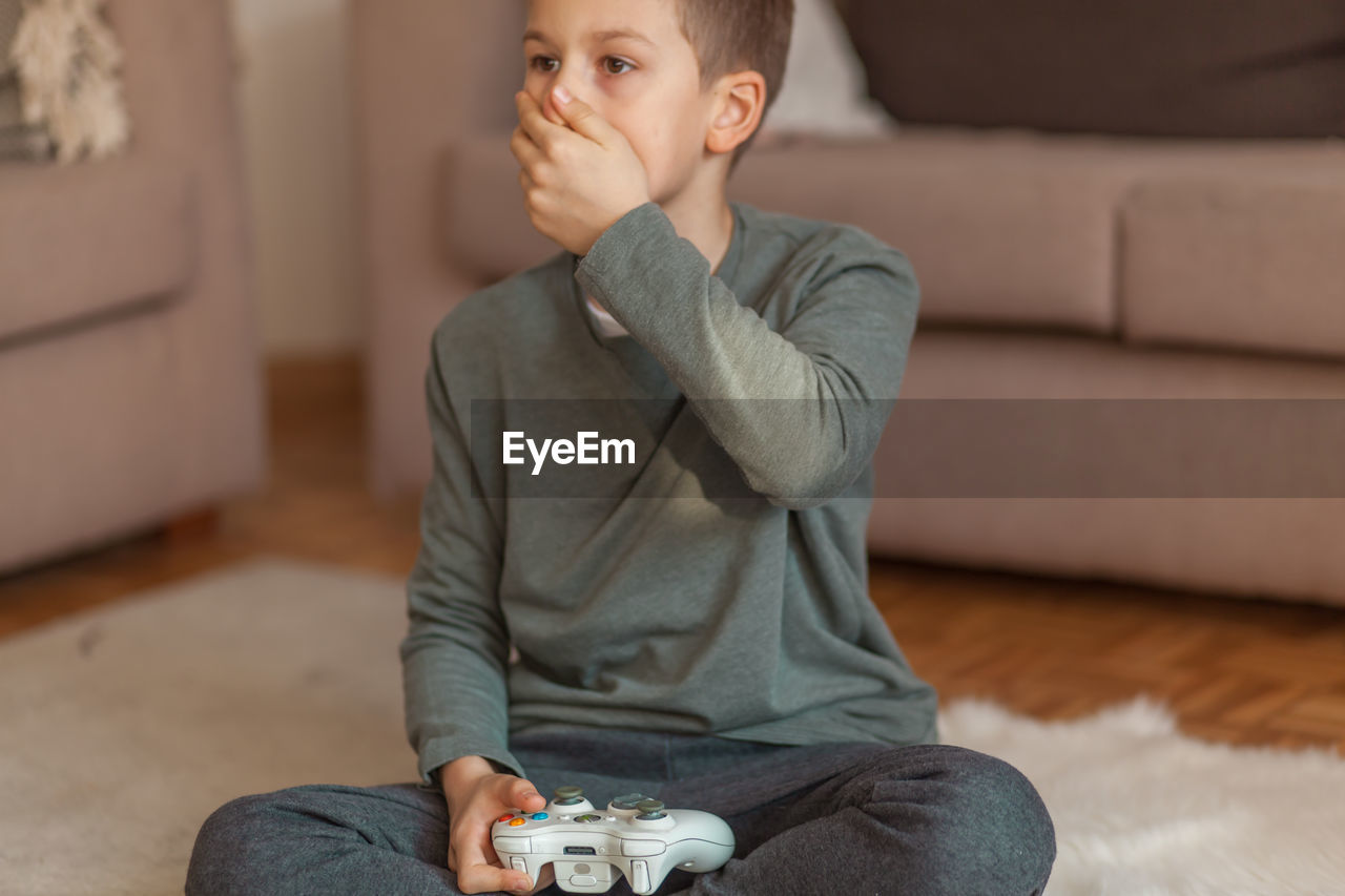 Boy with hand covering mouth holding video game remote control in living room at home 