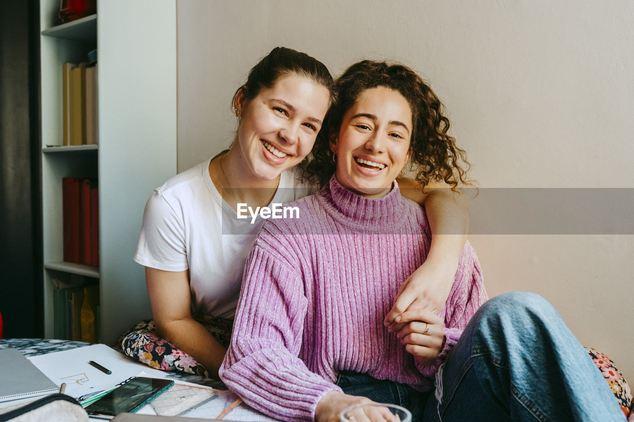 Portrait of happy young woman with arm around friend sitting at home