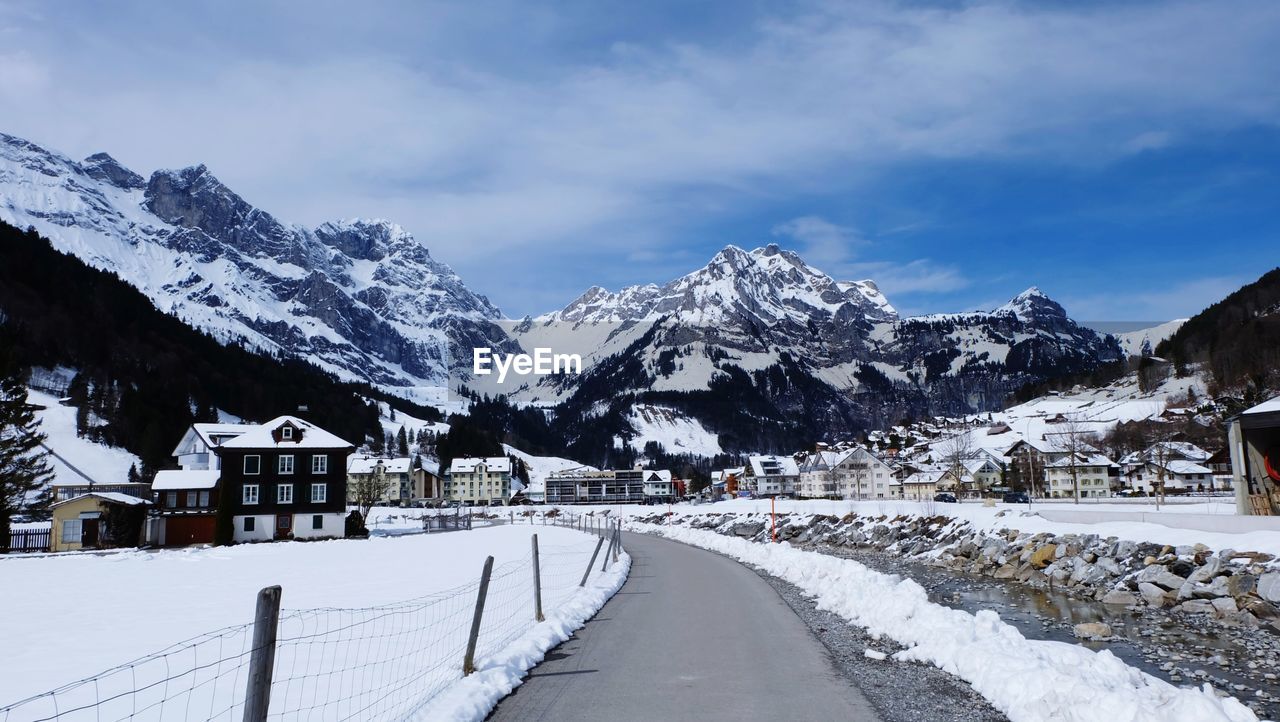 SNOW COVERED ROAD BY HOUSES AGAINST SKY