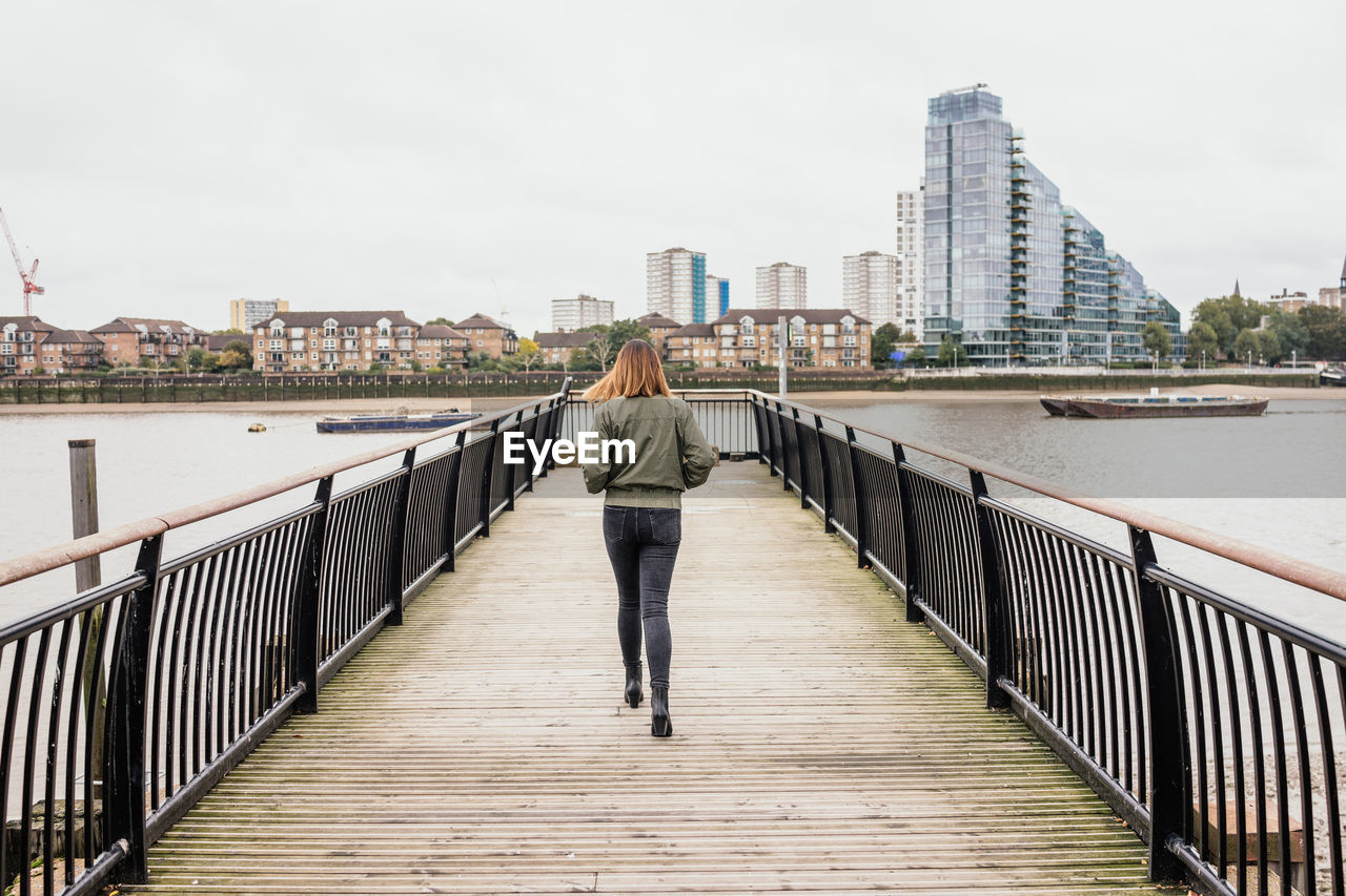 Rear view full length of woman walking over river on footbridge in city