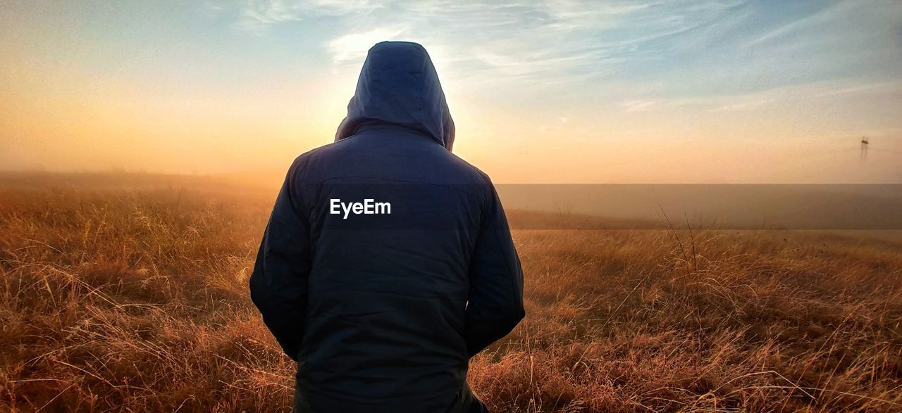 Rear view of man wearing hood standing on field against sky during sunset