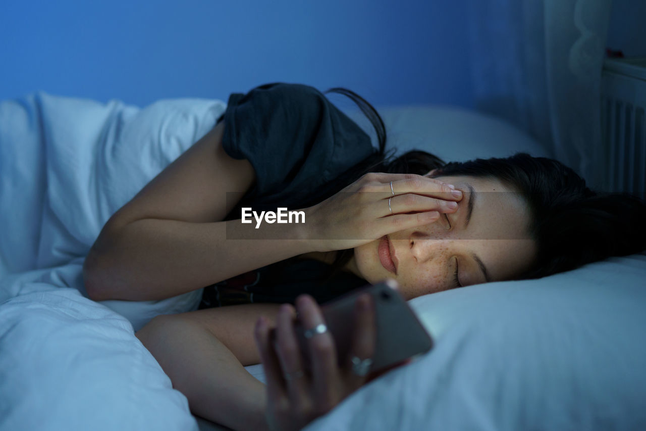 Exhausted woman looking at smartphone screen, lying in bed late at night. social media addiction
