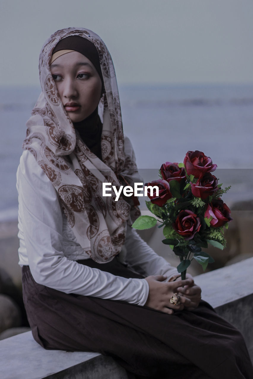 Thoughtful woman looking away while sitting with rose bouquet against sea