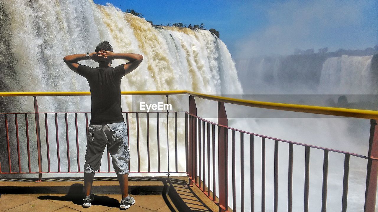 REAR VIEW OF MAN STANDING BY RAILING AGAINST WATER
