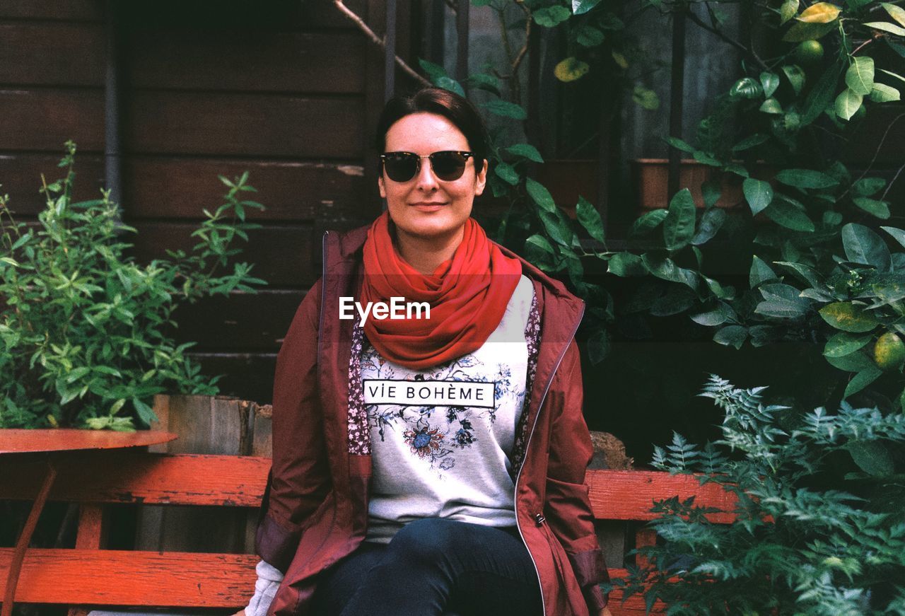 Portrait of smiling woman sitting on bench amidst plants at yard